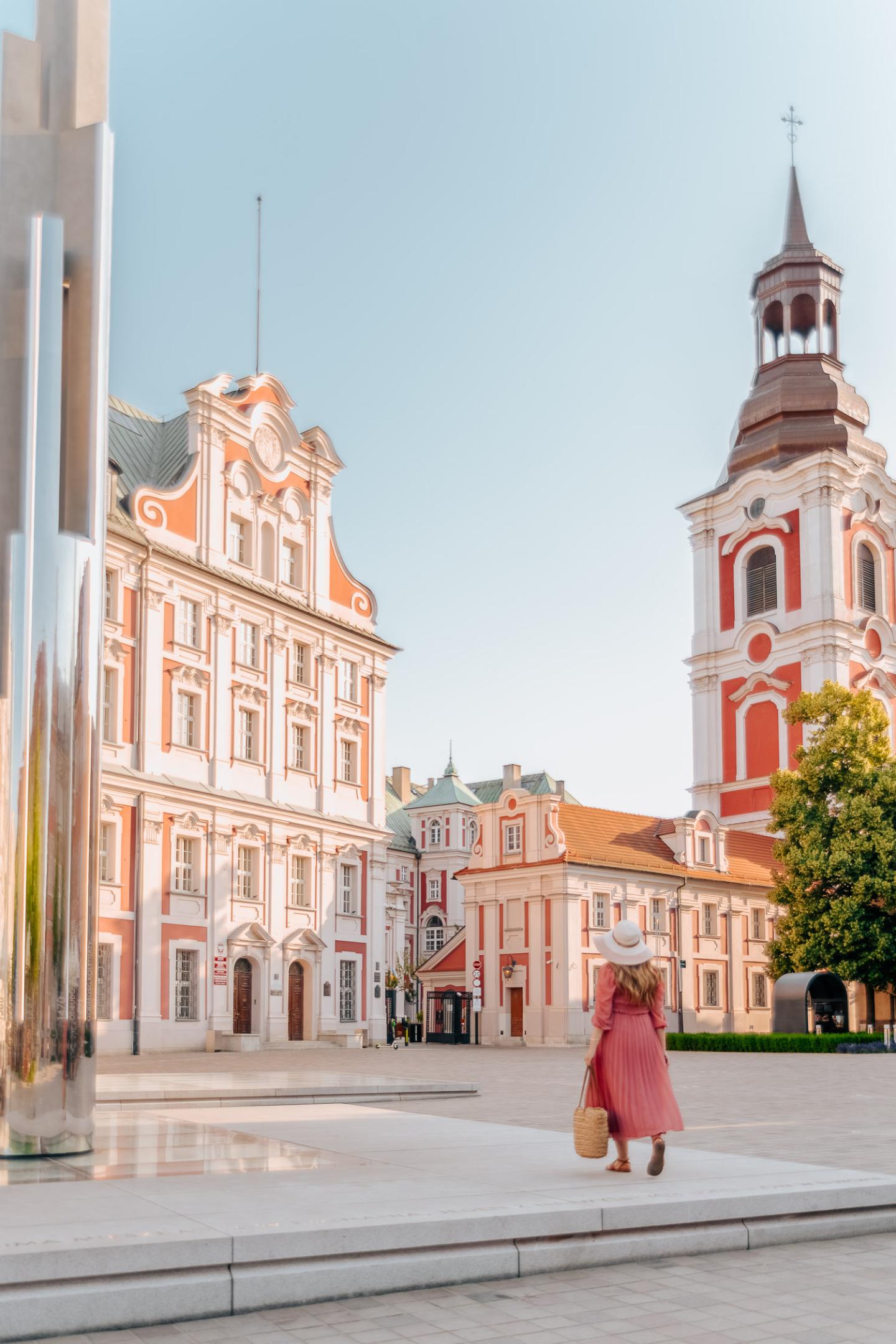 Woman in pink dress walking towards Poznań Fara in Poland.